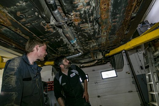 Two Auto Solve technicians inspecting a vehicle's exhaust and undercarriage on the lift